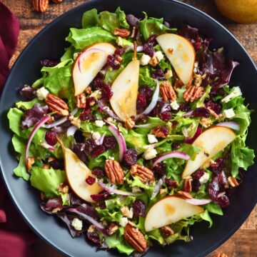 Overhead view of pear pecan fall salad with mixed greens, sliced pears, dried cranberries, toasted pecans, crumbled feta, and red onion in a black bowl on a rustic wood table.
