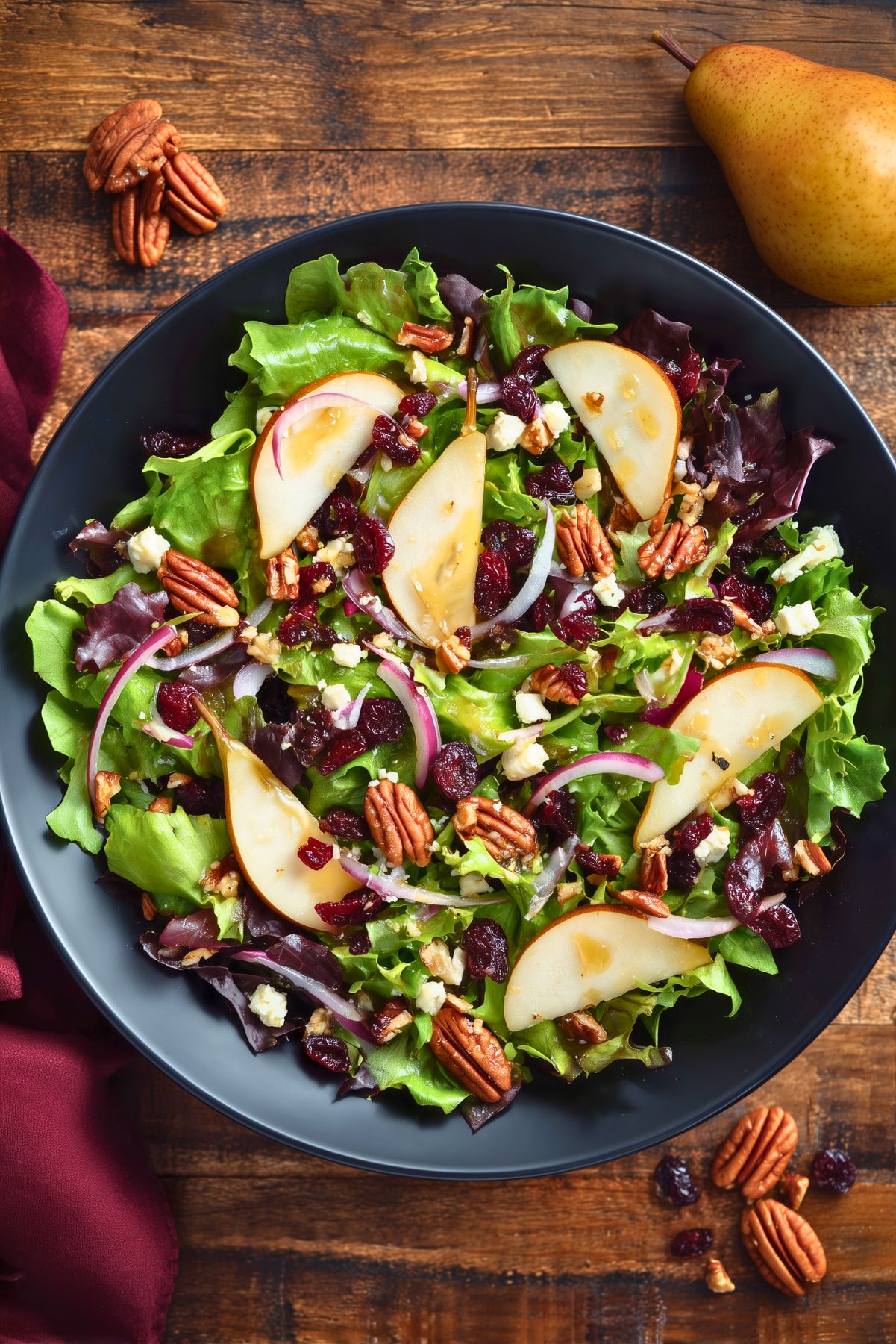 Overhead view of pear pecan fall salad with mixed greens, sliced pears, dried cranberries, toasted pecans, crumbled feta, and red onion in a black bowl on a rustic wood table.