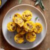 Overhead photo of pumpkin and turkey sausage egg bites piled on a beige plate over a rustic wood table, with a muffin pan and fresh sage leaves nearby.