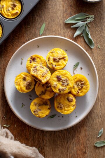 Overhead photo of pumpkin and turkey sausage egg bites piled on a beige plate over a rustic wood table, with a muffin pan and fresh sage leaves nearby.