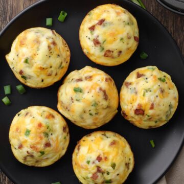 Six roast beef and cheddar egg bites with melted cheddar and green onions arranged on a black plate over a rustic wood table.