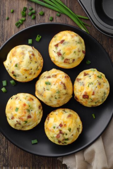 Six roast beef and cheddar egg bites with melted cheddar and green onions arranged on a black plate over a rustic wood table.