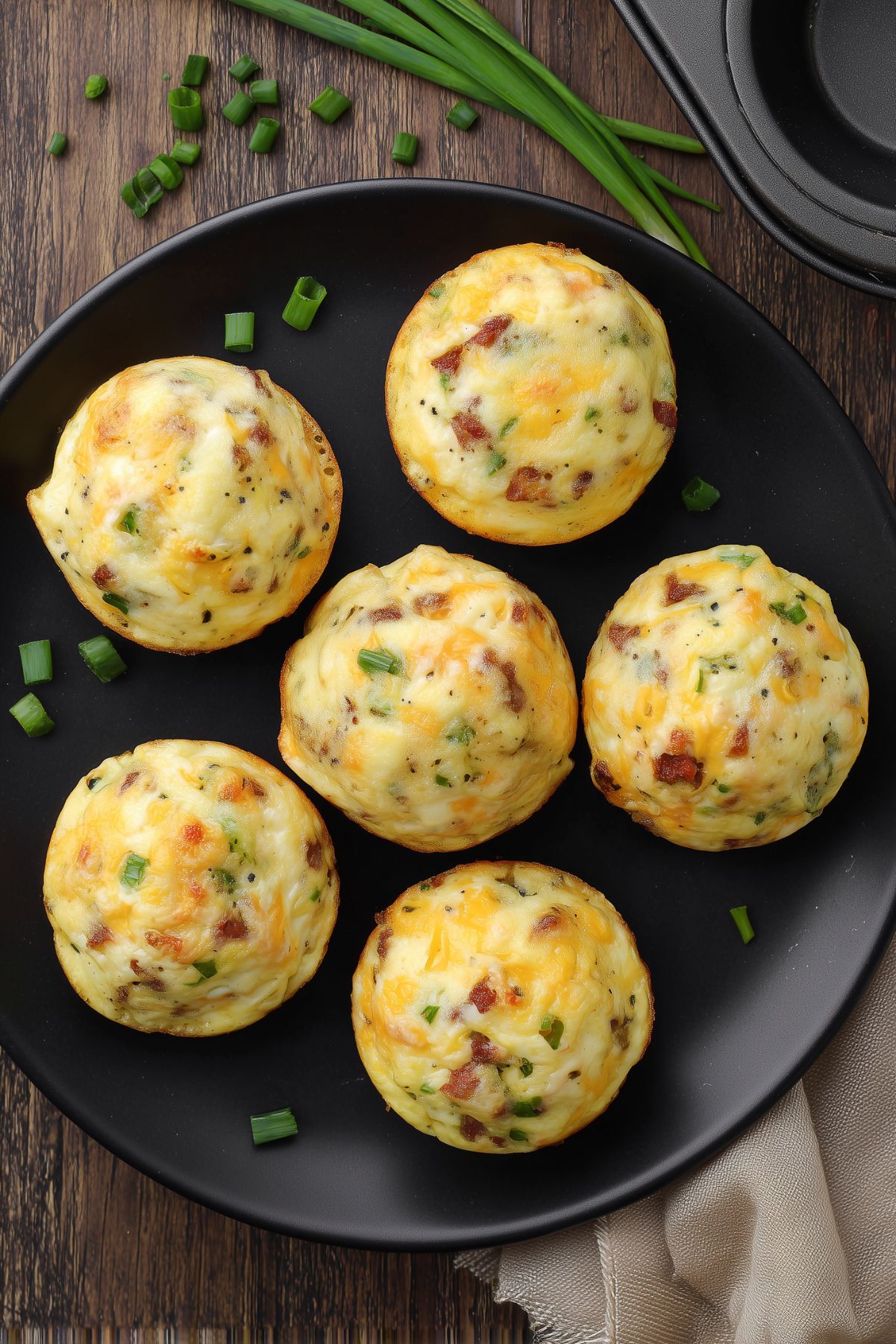 Six roast beef and cheddar egg bites with melted cheddar and green onions arranged on a black plate over a rustic wood table.