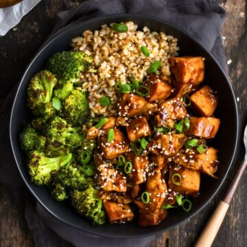 Overhead view of a sesame chicken sweet potato bowl with glazed chicken, roasted sweet potato cubes, broccoli, brown rice, sesame seeds, and green onions in a black bowl.