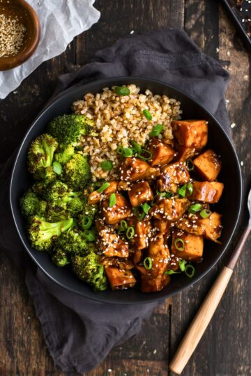 Overhead view of a sesame chicken sweet potato bowl with glazed chicken, roasted sweet potato cubes, broccoli, brown rice, sesame seeds, and green onions in a black bowl.