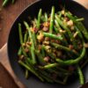 Overhead view of skillet green beans with bacon and garlic in a black bowl, with bright green beans, chopped bacon, sautéed onions, and seasoning on a rustic wooden table.