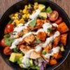 Overhead view of a Southwest chicken sweet potato bowl with roasted sweet potatoes, seasoned chicken, black beans, corn, avocado, cherry tomatoes, red onion, cilantro, and creamy sauce.