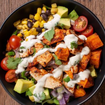 Overhead view of a Southwest chicken sweet potato bowl with roasted sweet potatoes, seasoned chicken, black beans, corn, avocado, cherry tomatoes, red onion, cilantro, and creamy sauce.