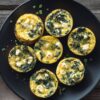 Overhead view of spinach feta egg bites with crumbled feta and green onions arranged on a matte black plate over a dark rustic wood table.