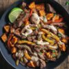 Overhead steak fajita sweet potato bowl with sliced steak, roasted sweet potatoes, sautéed bell peppers and onions, creamy fajita sauce, fresh herbs, and a lime wedge in a black bowl on a rustic wood table.