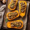 Overhead view of roasted butternut squash stuffed with quinoa and mushrooms on a parchment-lined baking sheet, surrounded by whole mushrooms, herbs, and a rustic wood background.