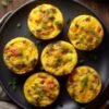 Six sweet potato breakfast hash egg bites with sausage, red bell pepper, cheddar, and green onions arranged on a black plate over a rustic wood table.
