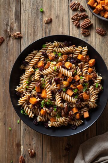 Overhead view of Sweet Potato Pecan Pasta Salad in a black bowl with rotini, roasted sweet potato cubes, dried cranberries, chopped pecans, and green onions on a rustic wood table.