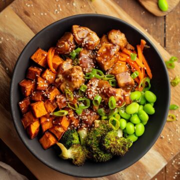 Teriyaki chicken sweet potato bowl with glazed chicken, roasted sweet potato cubes, broccoli, edamame, carrots, green onions, and sesame seeds in a black bowl on a rustic wooden table.