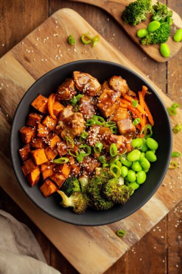 Teriyaki chicken sweet potato bowl with glazed chicken, roasted sweet potato cubes, broccoli, edamame, carrots, green onions, and sesame seeds in a black bowl on a rustic wooden table.