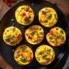 Overhead view of Tex-Mex egg bites with sausage, melted cheddar, red and green peppers, and green onions on a matte black plate over dark rustic wood.