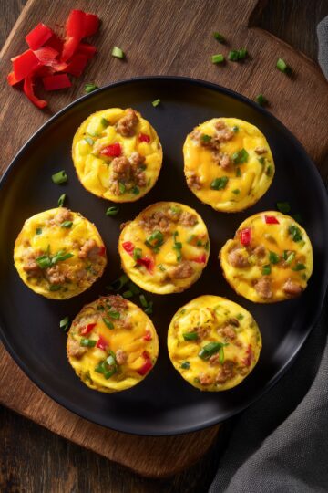 Overhead view of Tex-Mex egg bites with sausage, melted cheddar, red and green peppers, and green onions on a matte black plate over dark rustic wood.