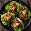 Overhead view of four Tex-Mex turkey lettuce wraps with ground turkey, black beans, corn, red peppers, cheddar cheese, sour cream, and green onions on a black plate.