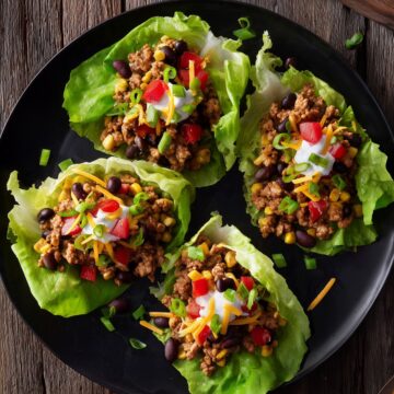 Overhead view of four Tex-Mex turkey lettuce wraps with ground turkey, black beans, corn, red peppers, cheddar cheese, sour cream, and green onions on a black plate.