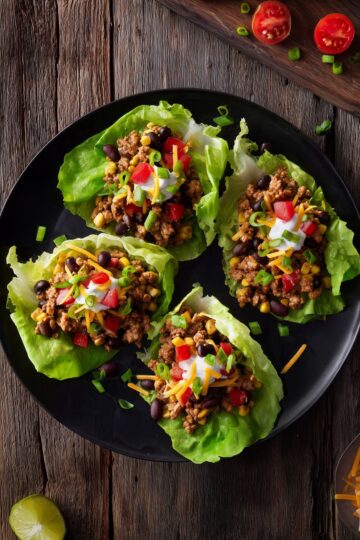 Overhead view of four Tex-Mex turkey lettuce wraps with ground turkey, black beans, corn, red peppers, cheddar cheese, sour cream, and green onions on a black plate.