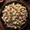 Overhead view of creamy Thanksgiving cranberry pecan pasta salad with rotini, apples, dried cranberries, pecans, celery, and parsley in a black bowl on a rustic wooden table.