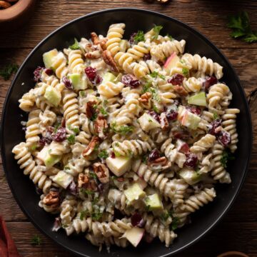 Overhead view of creamy Thanksgiving cranberry pecan pasta salad with rotini, apples, dried cranberries, pecans, celery, and parsley in a black bowl on a rustic wooden table.