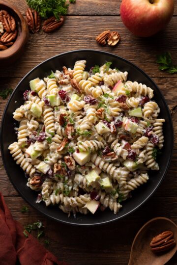 Overhead view of creamy Thanksgiving cranberry pecan pasta salad with rotini, apples, dried cranberries, pecans, celery, and parsley in a black bowl on a rustic wooden table.