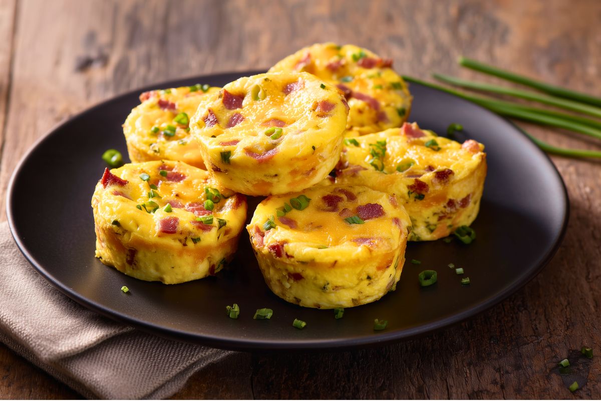 Turkey bacon and cheddar egg bites arranged on a black plate over a rustic wood table. The golden breakfast egg bites are filled with cheddar cheese, chopped turkey bacon, and green onions.