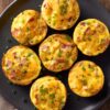 Overhead view of seven turkey bacon and cheddar egg bites on a black plate. The golden baked egg bites are topped with melted cheddar, pieces of turkey bacon, green onions, and black pepper.