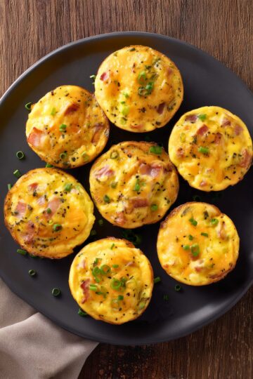 Overhead view of seven turkey bacon and cheddar egg bites on a black plate. The golden baked egg bites are topped with melted cheddar, pieces of turkey bacon, green onions, and black pepper.