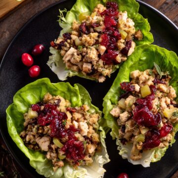 Overhead view of turkey cranberry stuffing lettuce wraps filled with chopped turkey, herb stuffing, celery, dried cranberries, and cranberry sauce on crisp butter lettuce, served on a black plate with fresh cranberries and herbs.