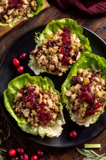 Overhead view of turkey cranberry stuffing lettuce wraps filled with chopped turkey, herb stuffing, celery, dried cranberries, and cranberry sauce on crisp butter lettuce, served on a black plate with fresh cranberries and herbs.