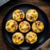 Overhead view of seven turkey sausage and apple egg bites on a matte black plate, with more egg bites in a muffin pan and sliced apples on a dark wood table.