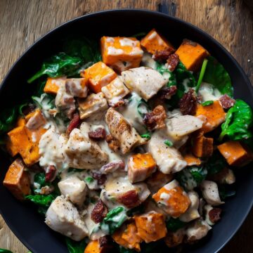 Overhead view of a Tuscan chicken sweet potato bowl with roasted sweet potatoes, chicken, spinach, bacon, and creamy sauce in a black bowl on a rustic wood table.