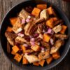 Close-up overhead view of an Apple Pecan Chicken Sweet Potato Bowl filled with seasoned chicken pieces, orange roasted sweet potato cubes, diced apples, dried cranberries, and pecan halves with a light sauce. Served in a matte black bowl on rustic wood with a red apple nearby.