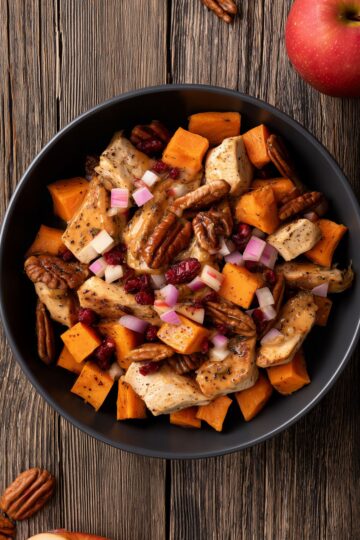 Close-up overhead view of an Apple Pecan Chicken Sweet Potato Bowl filled with seasoned chicken pieces, orange roasted sweet potato cubes, diced apples, dried cranberries, and pecan halves with a light sauce. Served in a matte black bowl on rustic wood with a red apple nearby.