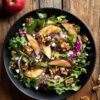 Overhead Apple Walnut Fall Salad with mixed greens, fresh apple slices, walnuts, dried cranberries, feta, red onion, and dressing in a black bowl on rustic wood.