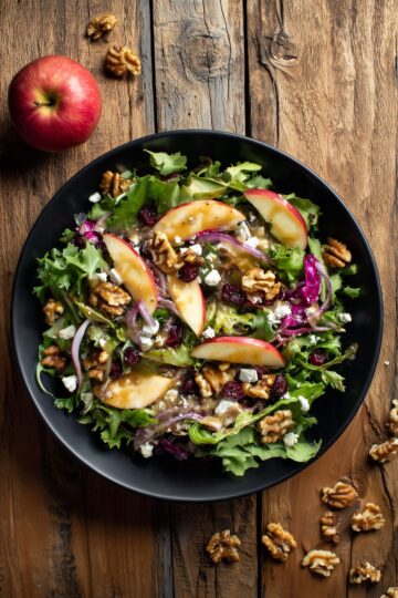 Overhead Apple Walnut Fall Salad with mixed greens, fresh apple slices, walnuts, dried cranberries, feta, red onion, and dressing in a black bowl on rustic wood.