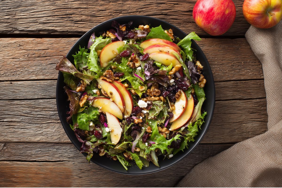 Apple Walnut Fall Salad with mixed greens, sliced red apples, walnuts, dried cranberries, feta, red onion, and dressing in a black bowl on a rustic wood table.