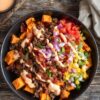 Close overhead view of a bacon cheeseburger sweet potato bowl filled with roasted sweet potato cubes, seasoned ground beef, crispy bacon pieces, shredded cheddar cheese, diced tomatoes, red onions, green onions, and creamy burger sauce. Served in a black bowl on rustic wood with extra sauce and pickles nearby.
