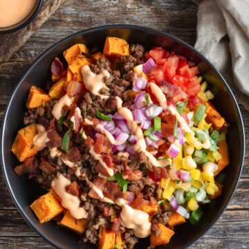Close overhead view of a bacon cheeseburger sweet potato bowl filled with roasted sweet potato cubes, seasoned ground beef, crispy bacon pieces, shredded cheddar cheese, diced tomatoes, red onions, green onions, and creamy burger sauce. Served in a black bowl on rustic wood with extra sauce and pickles nearby.