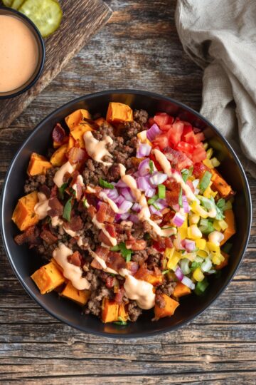 Close overhead view of a bacon cheeseburger sweet potato bowl filled with roasted sweet potato cubes, seasoned ground beef, crispy bacon pieces, shredded cheddar cheese, diced tomatoes, red onions, green onions, and creamy burger sauce. Served in a black bowl on rustic wood with extra sauce and pickles nearby.