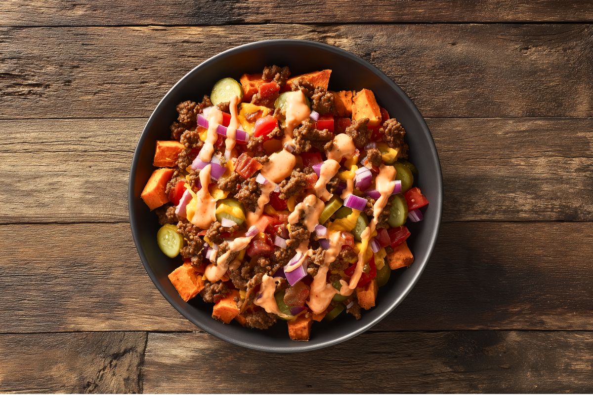 Overhead view of a bacon cheeseburger sweet potato bowl with roasted sweet potatoes, browned ground beef, crispy bacon, melted cheddar cheese, sliced pickles, diced tomatoes, red onions, and creamy burger sauce. Served in a matte black bowl on a rustic wooden table.