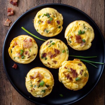 Overhead view of six Bacon Ranch Egg Bites with cheddar cheese, bacon, and green onions arranged on a black plate over a rustic wood table.