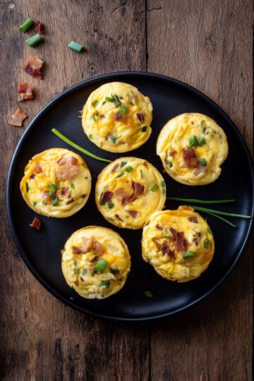 Overhead view of six Bacon Ranch Egg Bites with cheddar cheese, bacon, and green onions arranged on a black plate over a rustic wood table.