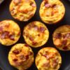 Overhead view of seven bacon and sweet potato egg bites with cheddar cheese on a matte black plate over a rustic wood table.