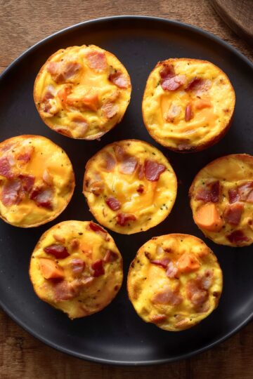 Overhead view of seven bacon and sweet potato egg bites with cheddar cheese on a matte black plate over a rustic wood table.