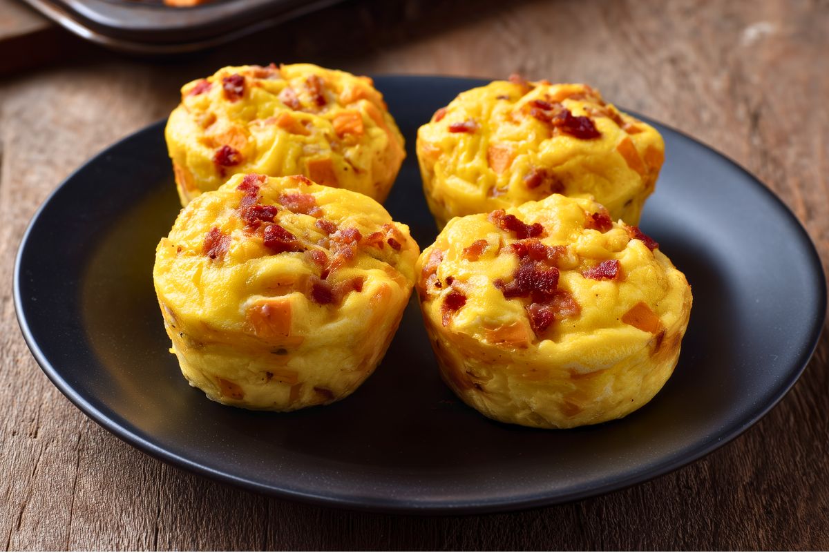 Four bacon and sweet potato egg bites with cheddar cheese arranged on a matte black plate over a rustic wood table.
