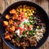 Overhead view of a Beef Burrito Sweet Potato Bowl in a matte black bowl with roasted sweet potato cubes, seasoned ground beef, black beans, rice, corn, diced tomatoes, sour cream, green onions, and cilantro. Served on a rustic wooden table.