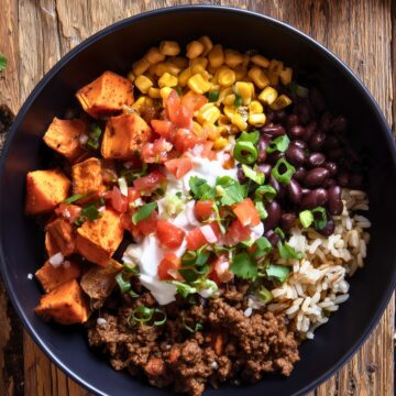Overhead view of a Beef Burrito Sweet Potato Bowl in a matte black bowl with roasted sweet potato cubes, seasoned ground beef, black beans, rice, corn, diced tomatoes, sour cream, green onions, and cilantro. Served on a rustic wooden table.